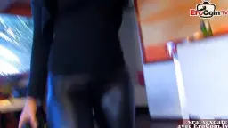 Blonde Frau mit tiefem Ausschnitt lächelt verführerisch in die Kamera, im Hintergrund der muskulöse Trainer