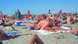 Nackte Paare auf bunten Handtüchern, Zuschauer stehen drumherum, heller Sandstrand