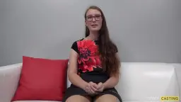 Schüchterne Brünette mit Brille und Blümchenkleid sitzt brav auf weißer Couch mit roten Kissen